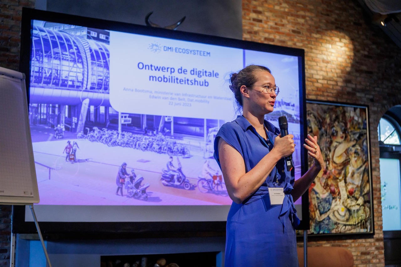 Foto van een vrouw die voor een videoscherm een zaal toespreekt