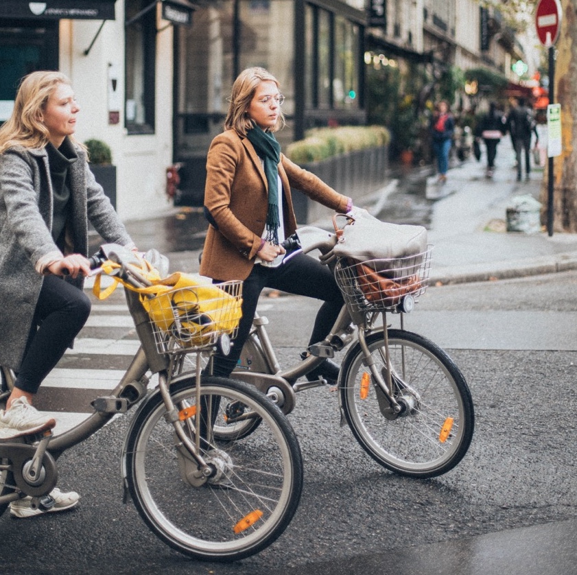Foto van 2 dames, vermoedelijk een moeder en dochter, op de fiets in een stad