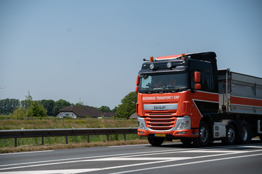 Foto van een rode vrachtwagen met aanhanger, rijdend op een provinciale weg met veel groen op de achtergrond