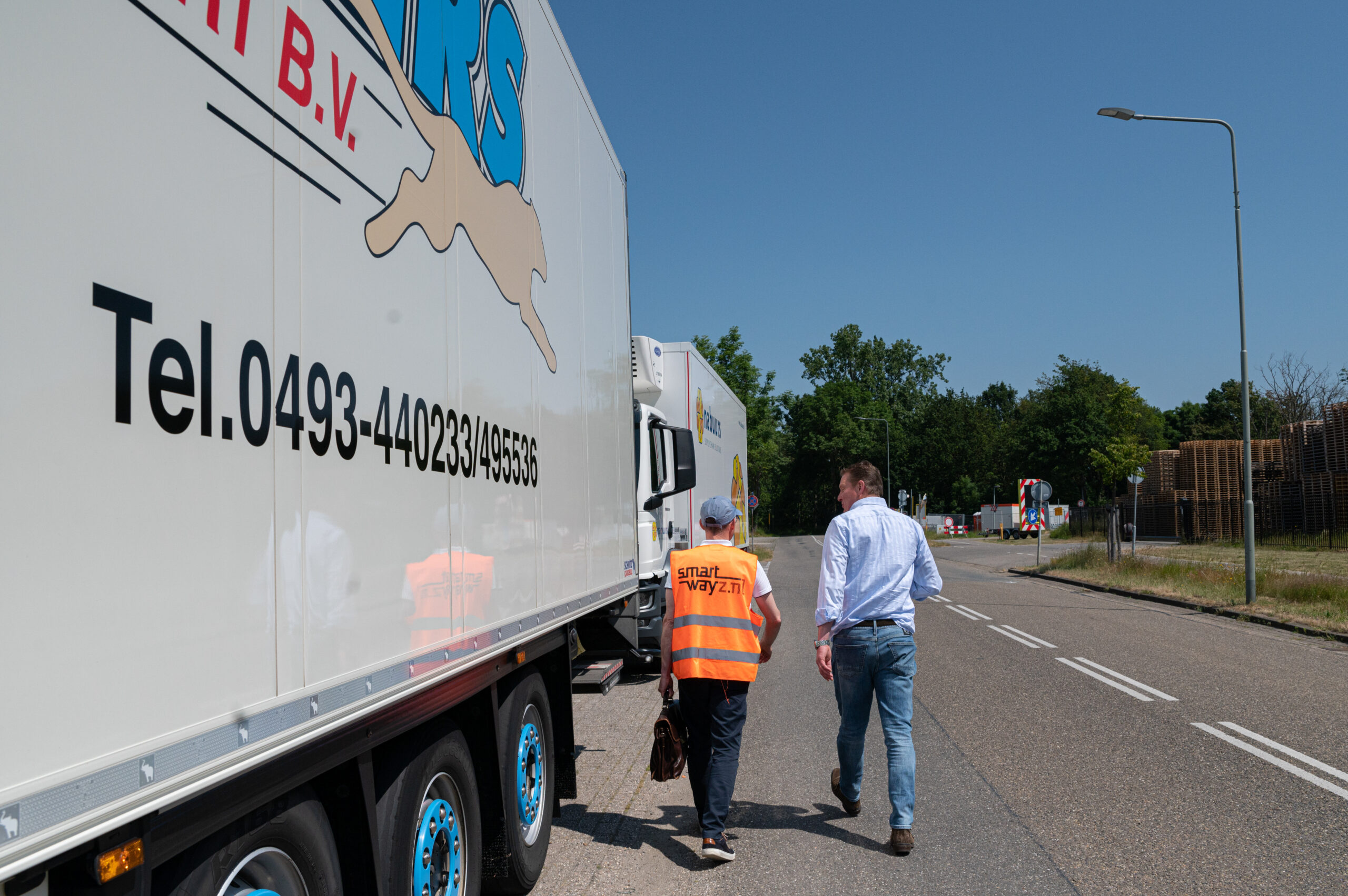 Foto. van twee heren, een met oranje veiligheidsvestje, lopen naast elkaar langs twee vrachtwagens